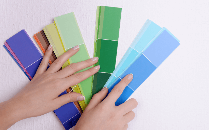 Image of a person holding different colour palette swatchespaint swatches and them holding it up to a wall.