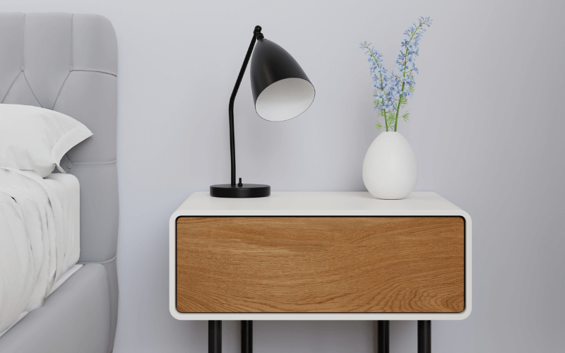 Image of a close-up shot on the side of a bedroom. A classy side drawer, decorated with simple items (a book or small stature), and a simple framed painting.