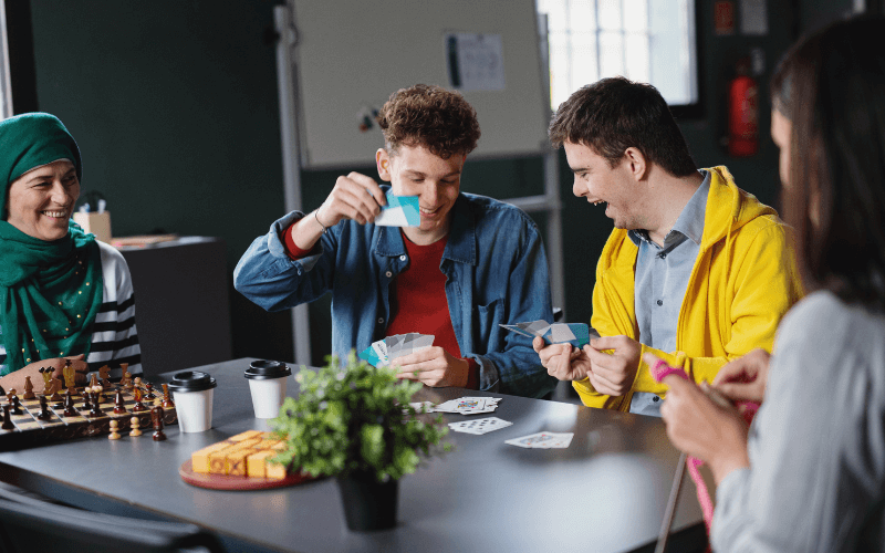 Image of a group of people gathered around a table with a board game in the centre. They all look like they’re having fun, laughing and smiling.